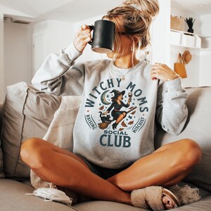 May include: A light grey sweatshirt featuring the text "Witchy Moms Social Club" and a witch graphic. The witch is on a broom, holding a wine glass. The sweatshirt is ash-coloured.