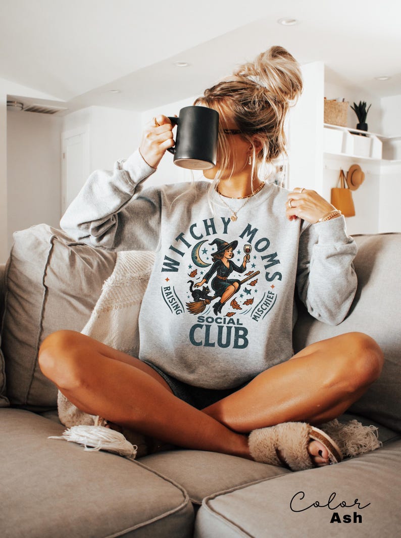 May include: A light grey sweatshirt featuring the text "Witchy Moms Social Club" and a witch graphic. The witch is on a broom, holding a wine glass. The sweatshirt is ash-coloured.