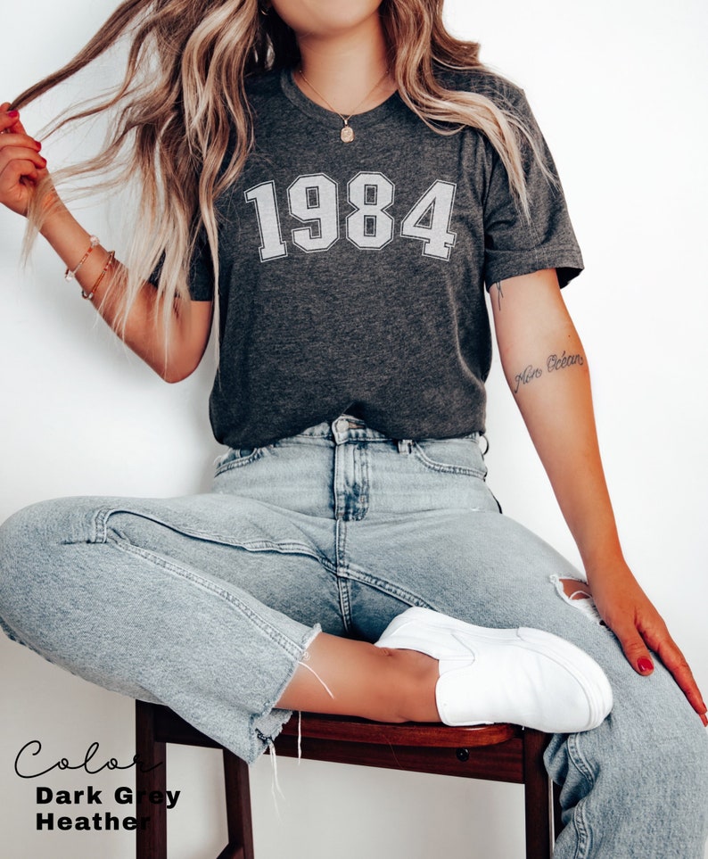 May include: A dark grey heather t-shirt with the year 1984 printed in white on the front. The shirt is being worn by a person with long blonde hair and blue jeans.