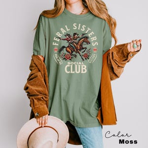 May include: A green t-shirt with a graphic of two women riding horses. The graphic says "Feral Sisters Social Club" with the words "Ride or Die" and "Sisters" on either side of the graphic.