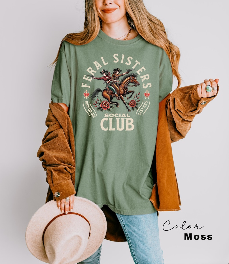 May include: A green t-shirt with a graphic of two women riding horses. The graphic says "Feral Sisters Social Club" with the words "Ride or Die" and "Sisters" on either side of the graphic.