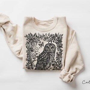 May include: A sand-colored sweatshirt featuring a detailed black and white illustration of an owl surrounded by floral and butterfly elements. The design is centered on the front of the long-sleeved garment.