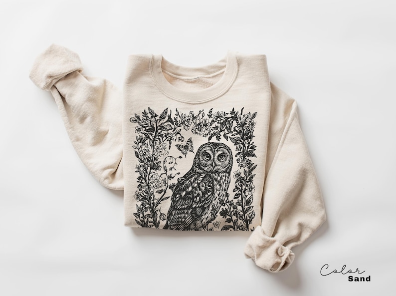May include: A sand-colored sweatshirt featuring a detailed black and white illustration of an owl surrounded by floral and butterfly elements. The design is centered on the front of the long-sleeved garment.