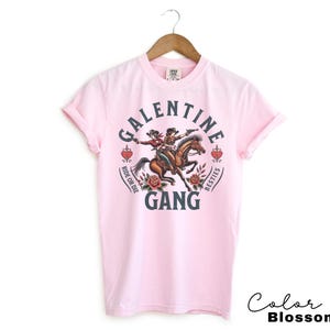 May include: Light pink t-shirt with a graphic that reads "Galentine Gang" with two women riding a horse. The design includes roses, hearts, and the phrases "Ride or Die" and "Besties". The words "Color Blossom" are written in cursive.