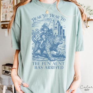 May include: A light blue t-shirt with a graphic featuring a knight riding a cat, a castle, and the text "Hear Ye! Hear Ye!" and "The Fun Aunt Has Arrived". The shirt has short sleeves and a crew neck.