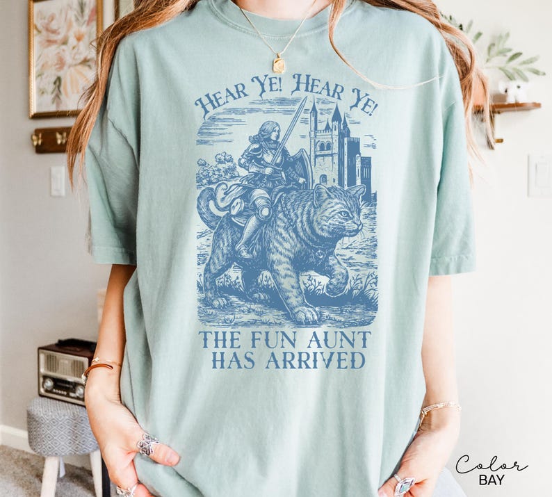 May include: A light blue t-shirt with a graphic featuring a knight riding a cat, a castle, and the text "Hear Ye! Hear Ye!" and "The Fun Aunt Has Arrived". The shirt has short sleeves and a crew neck.
