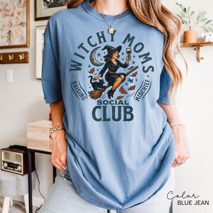 Puede incluir: Camiseta azul vaquero con un gr&aacute;fico que dice "Witchy Moms Social Club". El gr&aacute;fico presenta una bruja en una escoba con un gato negro, luna, estrellas y hojas de oto&ntilde;o. La camiseta es de corte holgado.