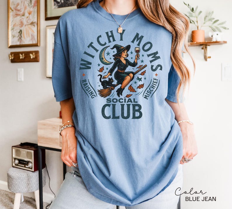 Puede incluir: Camiseta azul vaquero con un gr&aacute;fico que dice "Witchy Moms Social Club". El gr&aacute;fico presenta una bruja en una escoba con un gato negro, luna, estrellas y hojas de oto&ntilde;o. La camiseta es de corte holgado.