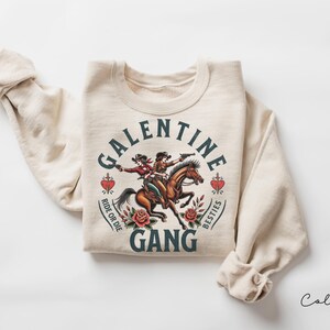 May include: Sand-colored sweatshirt with a graphic featuring two cowgirls riding a horse, guns drawn. Text reads "Galentine Gang" with "Ride or Die" and "Besties" around hearts and roses. A fun, western-themed design.