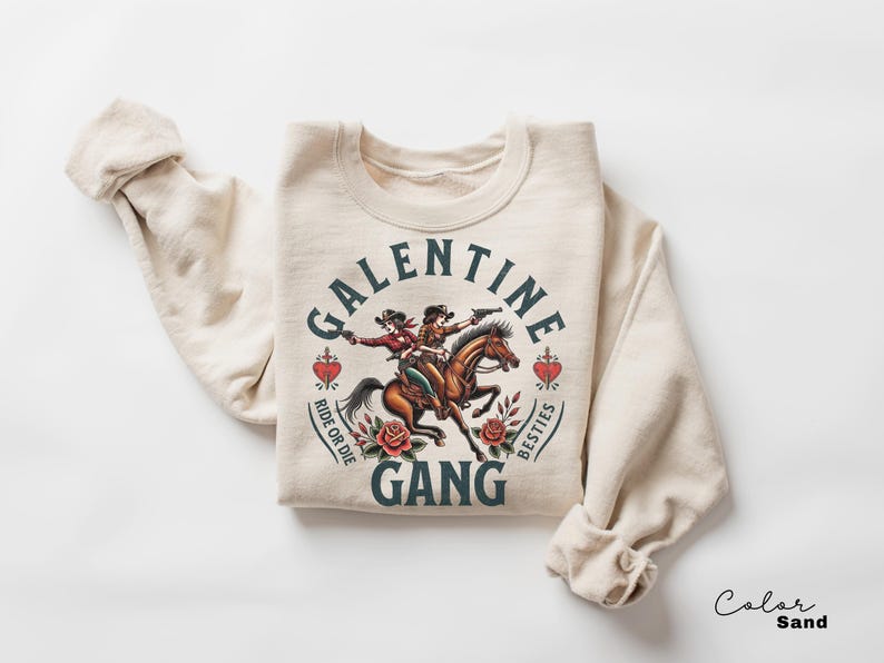 May include: Sand-colored sweatshirt with a graphic featuring two cowgirls riding a horse, guns drawn. Text reads "Galentine Gang" with "Ride or Die" and "Besties" around hearts and roses. A fun, western-themed design.