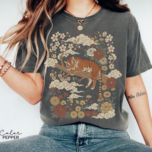 May include: Dark grey t-shirt featuring a tiger resting on a tree branch, surrounded by flowers and clouds. The design includes shades of brown, red, and yellow. The text "Color Pepper" is printed at the bottom of the image.