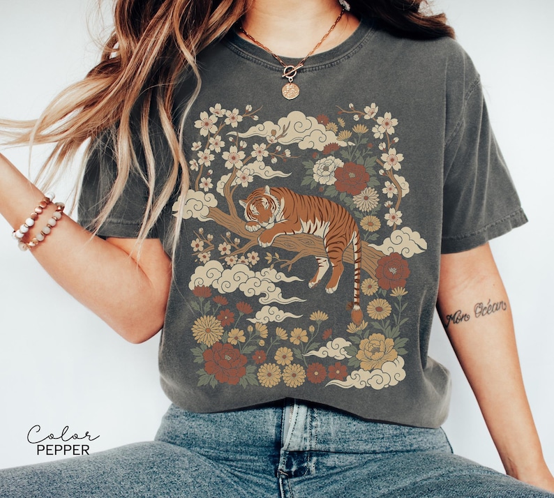 May include: Dark grey t-shirt featuring a tiger resting on a tree branch, surrounded by flowers and clouds. The design includes shades of brown, red, and yellow. The text "Color Pepper" is printed at the bottom of the image.