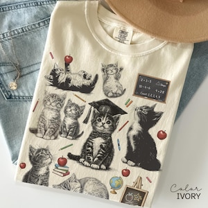 May include: Ivory-coloured t-shirt with a kitten-themed school design. The graphic features kittens, apples, pencils, and a chalkboard with sums. The shirt is made of a soft material.
