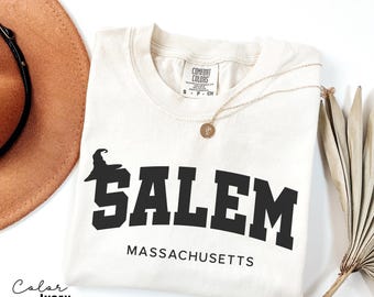 May include: Ivory-colored t-shirt with the word "SALEM" in bold black letters, a witch hat graphic, and "MASSACHUSETTS" below. The shirt has a crew neck and short sleeves. A gold necklace and a brown hat are visible.