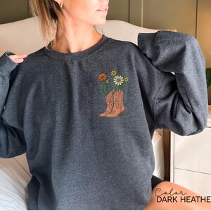 Puede incluir: Sudadera gris oscuro con un gr&aacute;fico de botas de vaquero llenas de flores. Las botas son de color marr&oacute;n claro y las flores incluyen flores naranjas y blancas. El texto "Color DARK HEATHER" est&aacute; en la parte inferior derecha.