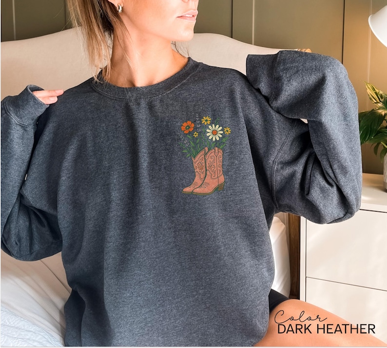 Puede incluir: Sudadera gris oscuro con un gr&aacute;fico de botas de vaquero llenas de flores. Las botas son de color marr&oacute;n claro y las flores incluyen flores naranjas y blancas. El texto "Color DARK HEATHER" est&aacute; en la parte inferior derecha.