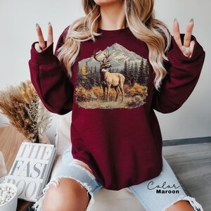 May include: Maroon sweatshirt featuring a detailed graphic of a deer in a mountain landscape. The design includes a deer with large antlers, trees, and a mountain range. The text "Color Maroon" is visible on the bottom right.