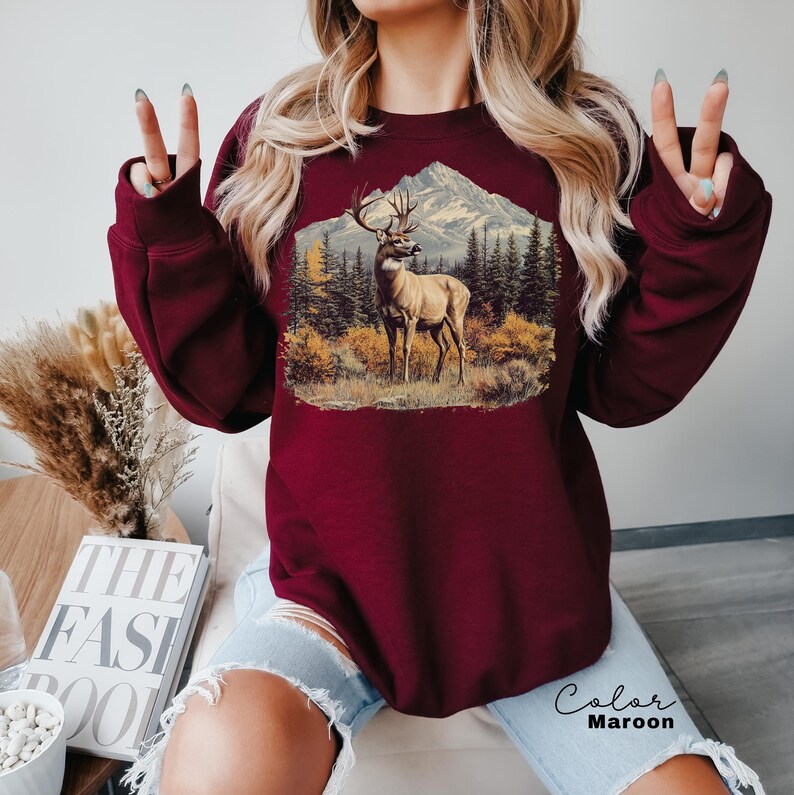 May include: Maroon sweatshirt featuring a detailed graphic of a deer in a mountain landscape. The design includes a deer with large antlers, trees, and a mountain range. The text "Color Maroon" is visible on the bottom right.