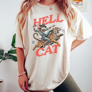 Könnte beinhalten: Ein cremefarbenes T-Shirt mit einem Aufdruck einer Frau, die auf einem Tiger reitet. Der Aufdruck ist in Rot, Blau und Schwarz gehalten. Der Text "HELL CAT" ist in Rot auf das Shirt gedruckt.