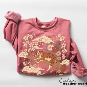 May include: Heather scarlet red sweatshirt with a tiger graphic. The design features a tiger resting on a tree branch, surrounded by flowers and clouds. The text "Color Heather Scarlet Red" is at the bottom.