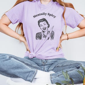 Puede incluir: Una camiseta lavanda con una ilustraci&oacute;n de estilo retro de una mujer sosteniendo un cuchillo y el texto "Mentally Spicy".