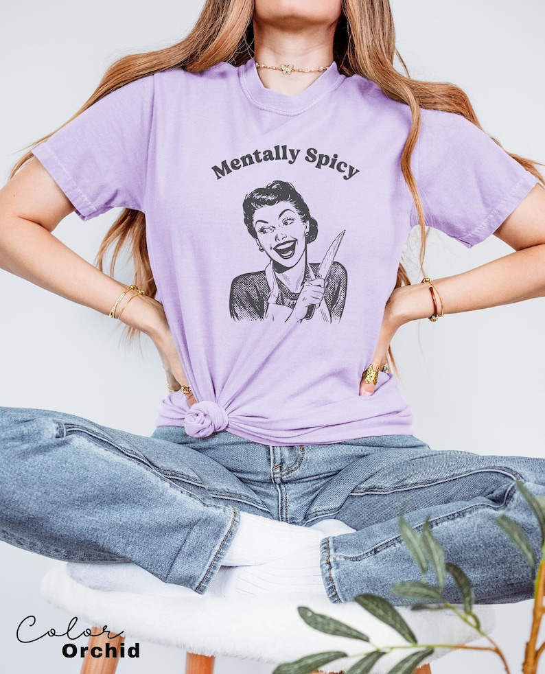 Puede incluir: Una camiseta lavanda con una ilustraci&oacute;n de estilo retro de una mujer sosteniendo un cuchillo y el texto "Mentally Spicy".
