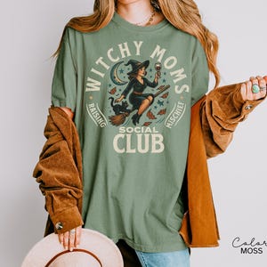 Puede incluir: Camiseta verde salvia con un dise&ntilde;o de una bruja en una escoba, un gato negro y las palabras "Witchy Moms Social Club". El dise&ntilde;o incluye una luna creciente, estrellas y hojas de oto&ntilde;o. La camiseta es de corte holgado.