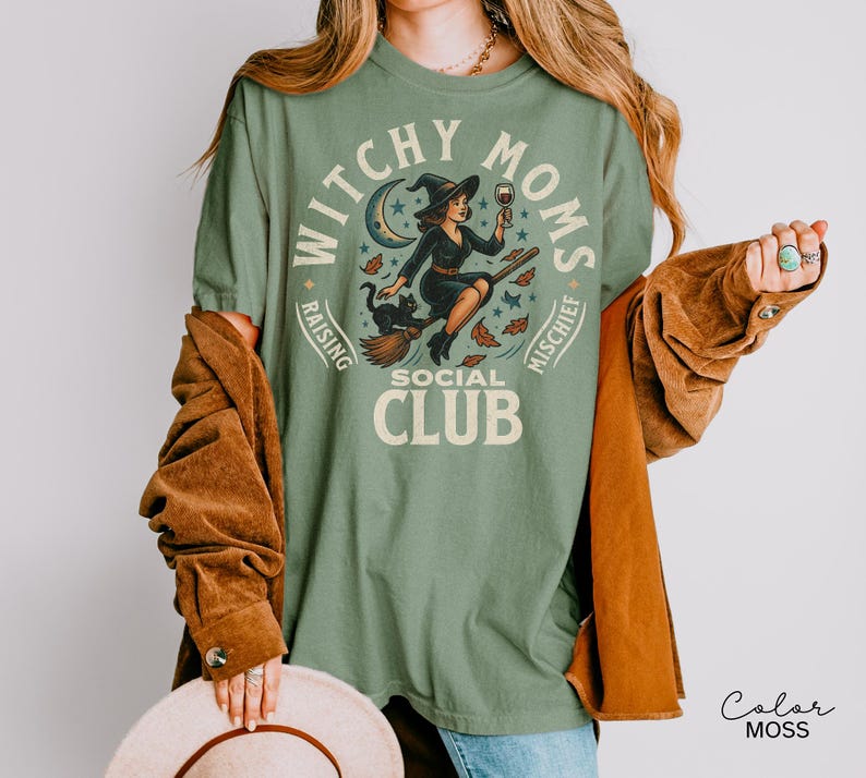 Puede incluir: Camiseta verde salvia con un dise&ntilde;o de una bruja en una escoba, un gato negro y las palabras "Witchy Moms Social Club". El dise&ntilde;o incluye una luna creciente, estrellas y hojas de oto&ntilde;o. La camiseta es de corte holgado.
