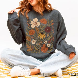 Puede incluir: Una sudadera de manga larga en color gris jaspeado oscuro con un estampado gr&aacute;fico central de varias flores y hojas en tonos oto&ntilde;ales de crema, naranja, &oacute;xido y amarillo. El texto "Color Dark Heather" est&aacute; impreso en la esquina inferior derecha.