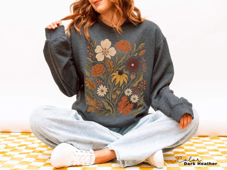 Puede incluir: Una sudadera de manga larga en color gris jaspeado oscuro con un estampado gr&aacute;fico central de varias flores y hojas en tonos oto&ntilde;ales de crema, naranja, &oacute;xido y amarillo. El texto "Color Dark Heather" est&aacute; impreso en la esquina inferior derecha.