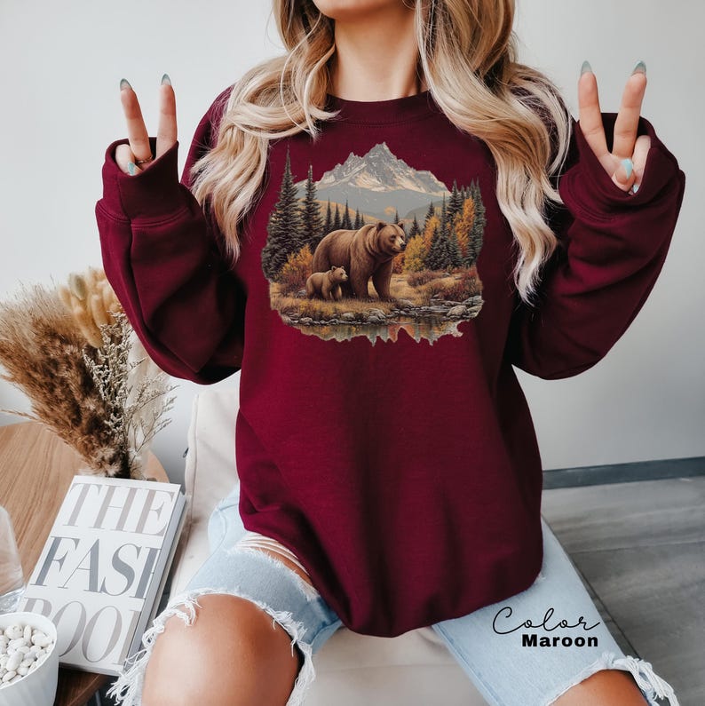 May include: Maroon sweatshirt featuring a nature scene with a bear and cub near a mountain and trees. The text "Color Maroon" is written in cursive at the bottom. The sweatshirt is a solid color and has long sleeves.