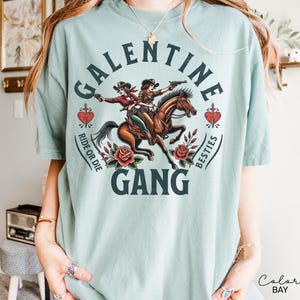 May include: A light blue t-shirt featuring a graphic of two cowgirls riding a horse, with the text "GALENTINE GANG" and "RIDE OR DIE BESTIES". The design includes roses and hearts.