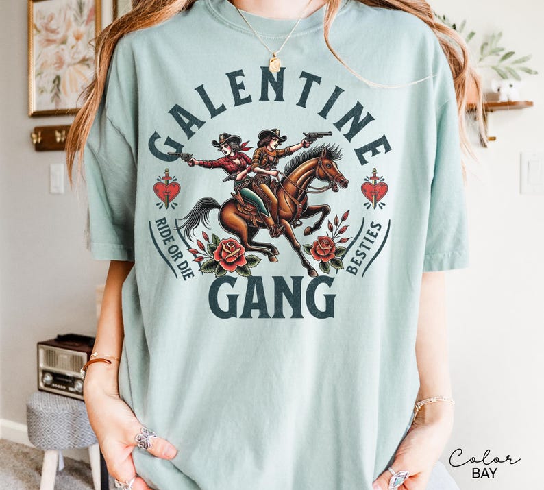 May include: A light blue t-shirt featuring a graphic of two cowgirls riding a horse, with the text "GALENTINE GANG" and "RIDE OR DIE BESTIES". The design includes roses and hearts.