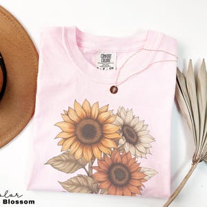 May include: A pink t-shirt featuring a graphic of three sunflowers in shades of yellow, orange, and brown. The shirt has the text "Comfort Colors" on the tag.