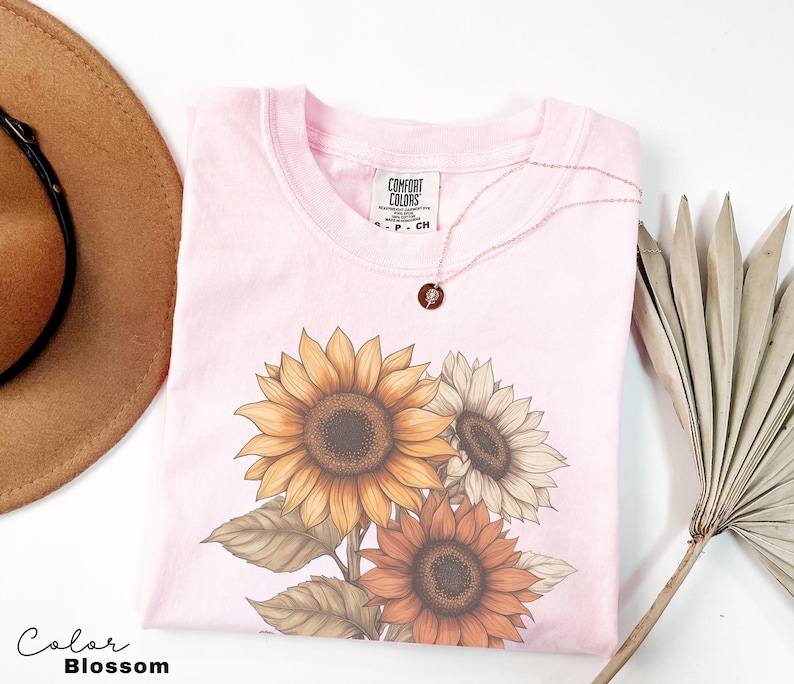 May include: A pink t-shirt featuring a graphic of three sunflowers in shades of yellow, orange, and brown. The shirt has the text "Comfort Colors" on the tag.