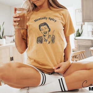 Puede incluir: Una mujer lleva una camiseta amarilla con el texto "Mentally Spicy" y una ilustraci&oacute;n retro de una mujer sosteniendo un cuchillo. Est&aacute; sentada en una superficie blanca con las piernas cruzadas.