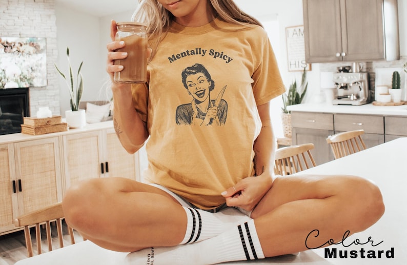 Puede incluir: Una mujer lleva una camiseta amarilla con el texto "Mentally Spicy" y una ilustraci&oacute;n retro de una mujer sosteniendo un cuchillo. Est&aacute; sentada en una superficie blanca con las piernas cruzadas.