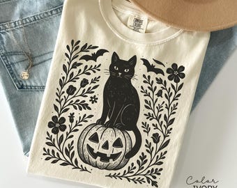 Könnte beinhalten: Elfenbeinfarbenes T-Shirt mit einer schwarzen Katze auf einem Kürbis. Das Design umfasst Fledermäuse, Blumen und Blätter. Der Text "Color IVORY" ist sichtbar. Das Shirt wird mit einer blauen Jeans und einer Halskette gezeigt.