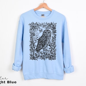 May include: Light blue sweatshirt featuring a black and white owl illustration within a floral frame. The design includes a butterfly. The text "Color Light Blue" is at the bottom left. The sweatshirt is hanging on a wooden hanger.