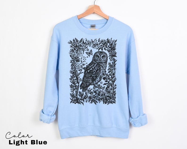 May include: Light blue sweatshirt featuring a black and white owl illustration within a floral frame. The design includes a butterfly. The text "Color Light Blue" is at the bottom left. The sweatshirt is hanging on a wooden hanger.