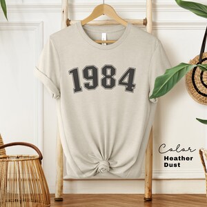 May include: A heather dust colored t-shirt with the number 1984 printed on the front in black.
