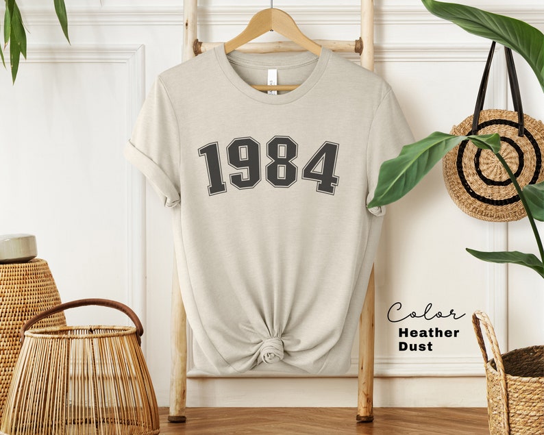 May include: A heather dust colored t-shirt with the number 1984 printed on the front in black.