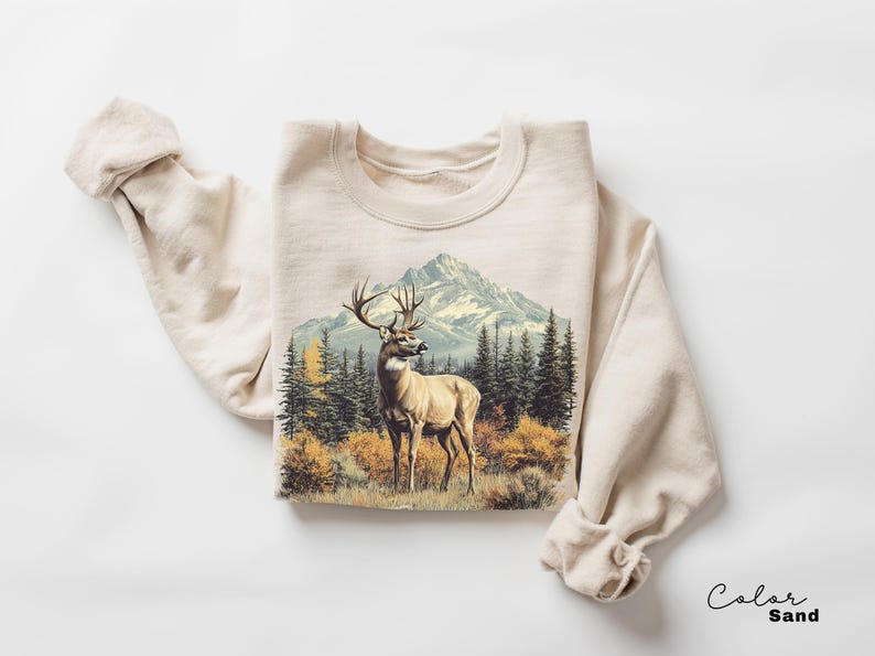 May include: A sand-colored sweatshirt featuring a detailed graphic of a deer in a forest with a mountain backdrop. The deer has large antlers and is standing in a field of grass and bushes. The sweatshirt is folded with the sleeves rolled up.