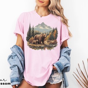 May include: A light pink t-shirt featuring a nature scene with a bear and cub in a forest setting, with mountains in the background. The text "Color Blossom" is at the bottom left.