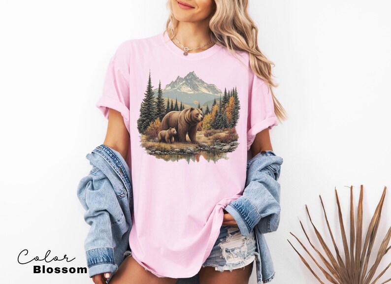 May include: A light pink t-shirt featuring a nature scene with a bear and cub in a forest setting, with mountains in the background. The text "Color Blossom" is at the bottom left.