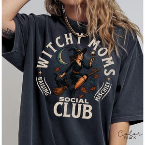 Puede incluir: Camiseta negra con un dise&ntilde;o circular. Presenta una bruja en una escoba sosteniendo una copa de vino, un gato negro y el texto "Witchy Moms Social Club". El dise&ntilde;o incluye una luna creciente y hojas de oto&ntilde;o.