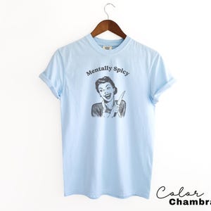 Puede incluir: Camiseta azul claro con un gr&aacute;fico de una mujer sosteniendo un cuchillo y el texto "Mentally Spicy".