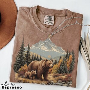May include: Espresso-colored t-shirt featuring a graphic of a bear and cub in a forest with mountains in the background. The shirt has a crew neck and short sleeves. A silver necklace with a pendant is draped on the shirt.