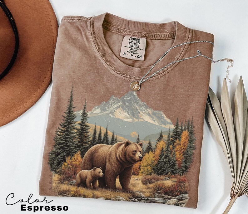 May include: Espresso-colored t-shirt featuring a graphic of a bear and cub in a forest with mountains in the background. The shirt has a crew neck and short sleeves. A silver necklace with a pendant is draped on the shirt.