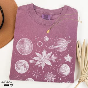 Puede incluir: Una camiseta de color malva con un estampado gr&aacute;fico blanco de planetas, estrellas y flores. El texto "Comfort Colors" est&aacute; impreso en la camiseta.